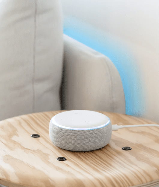 Wyze Light Strip lighting up the side of a couch and a voice assistant device placed on a table. 