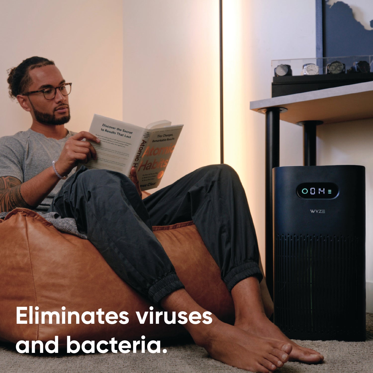 Man reading a book nearby a watch collection and Wyze Air Purifier. White text overlay that says "eliminates viruses and bacteria."